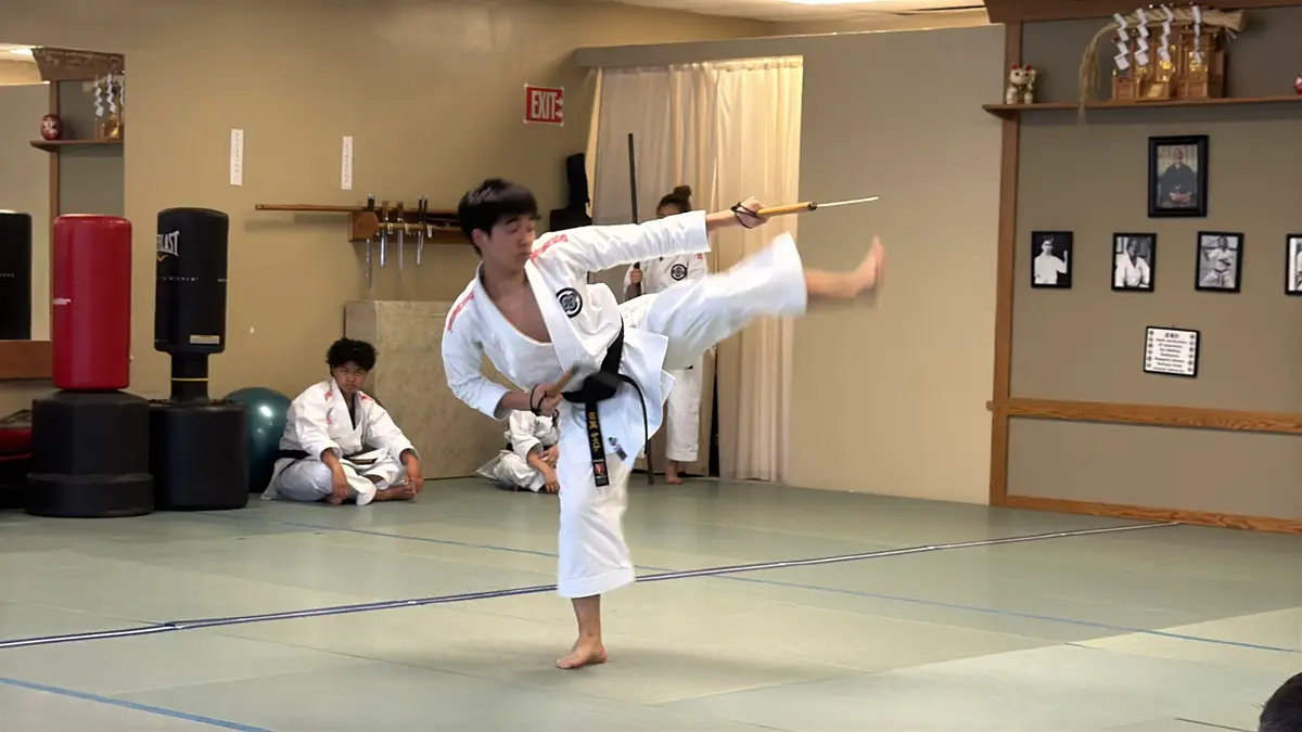 Black belt demonstrating a high kick while holding training weapons